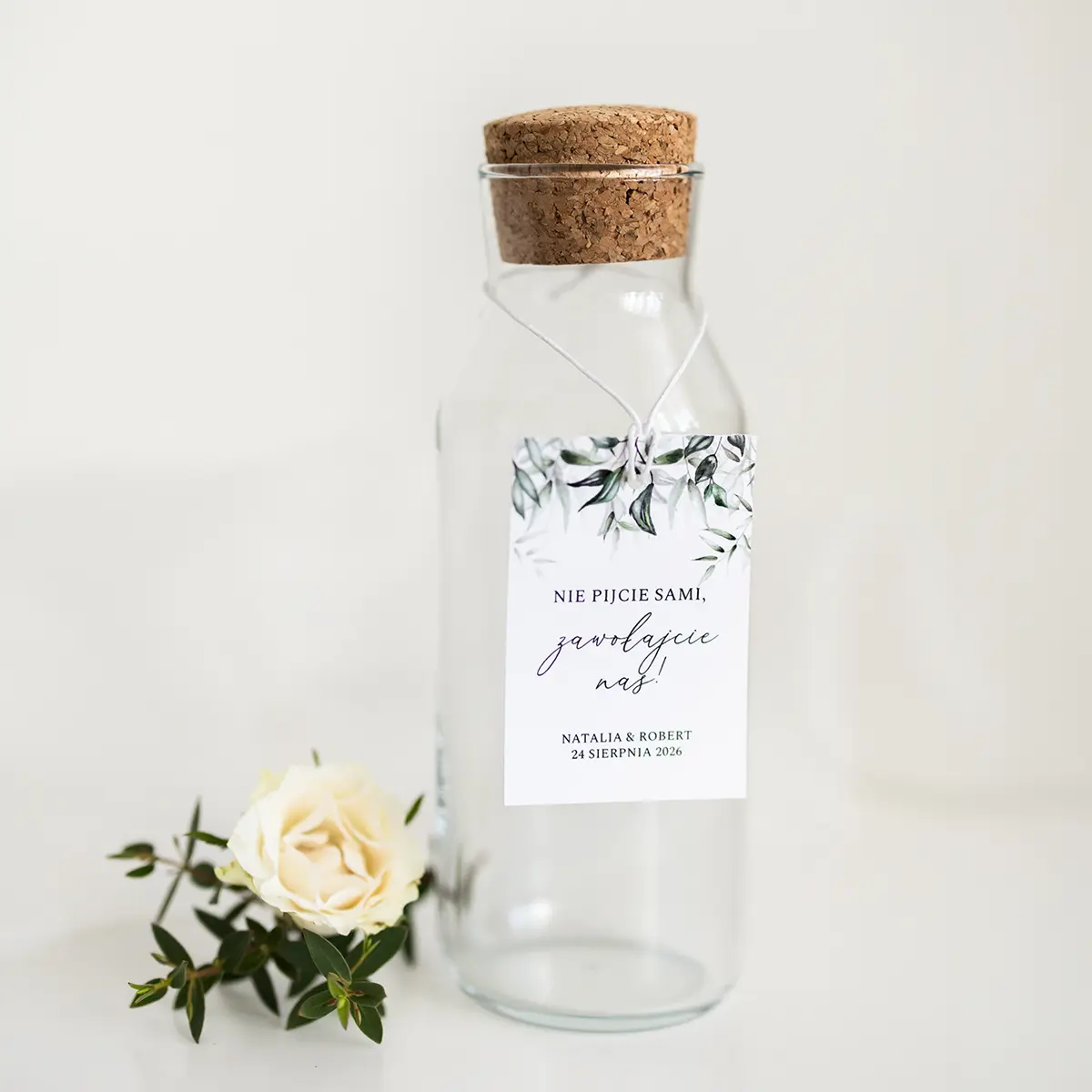 Elegancka zawieszka na butelkę z zielonym motywem roślinnym, przewiązana sznurkiem, na tle białej powierzchni z dekoracjami.