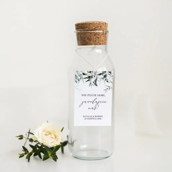 Zawieszka na alkohol z zielonymi liśćmi, zawieszona na przezroczystej butelce z korkiem, w towarzystwie białej róży.