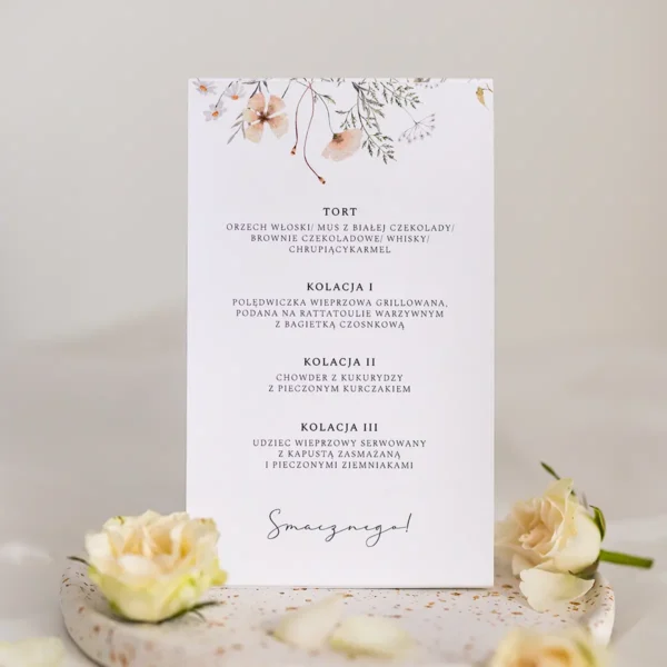 Tył stojącego menu ślubnego z kwiatowym nadrukiem, ułożonego na tle białych płatków róż.