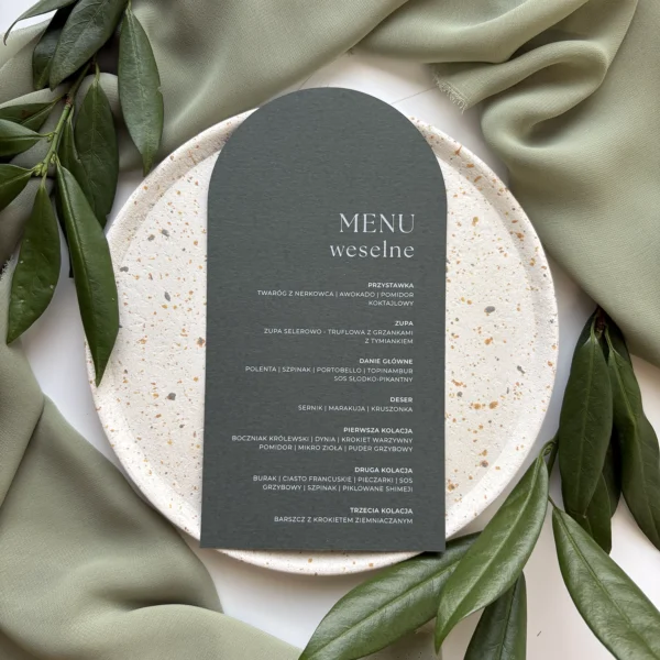 Menu weselne Jungle w butelkowej zieleni z eleganckim nadrukiem, ułożone na jasnym talerzu i otoczone zielonymi liśćmi.