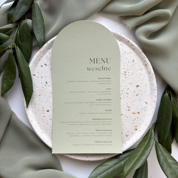 Menu weselne z kolekcji Jungle w odcieniu jasnej zieleni, ułożone na talerzu z beżowymi akcentami i zielonymi liśćmi w tle.