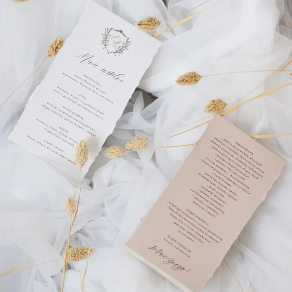 Beżowa i biała karta menu weselnego z kolekcji Róża z dekoracyjnymi, szarpanymi brzegami, ustawione na tle z tiulu i suchych traw.