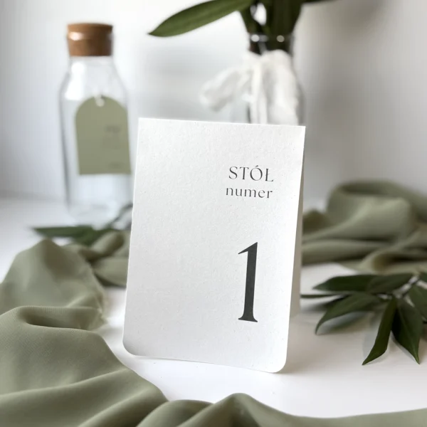 Numer stołu Jungle w kolorze złamanej bieli, ustawiony na tle butelek i zielonych tkanin w naturalnym stylu.