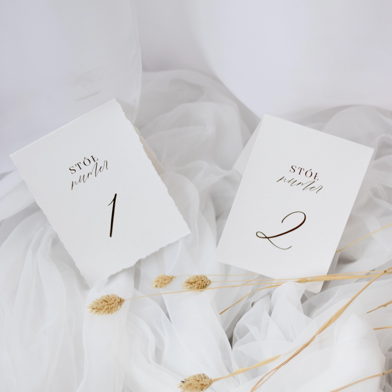 Dwa warianty numerów stołów z kolekcji Róża: jeden na gładkim białym papierze, drugi z postrzępionym brzegiem. Elegancki minimalizm i delikatna typografia.