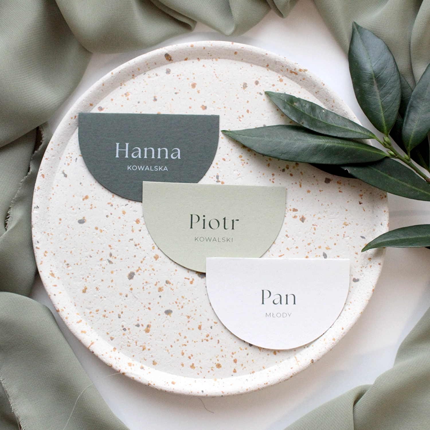 Półokrągłe winietki ślubne z kolekcji Jungle w odcieniach zieleni i złamanej bieli, ułożone na dekoracyjnym talerzu.