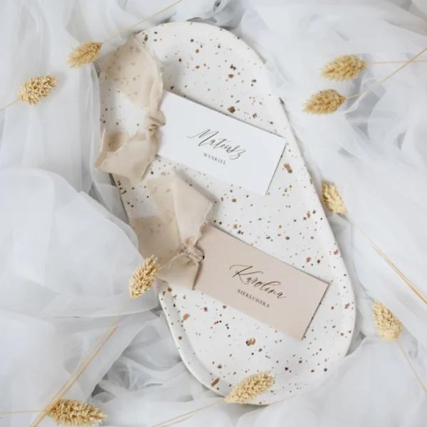 Cztery winietki z nazwiskami gości w różnych odcieniach beżu i bieli, ułożone na białym tle z suszonymi trawami.