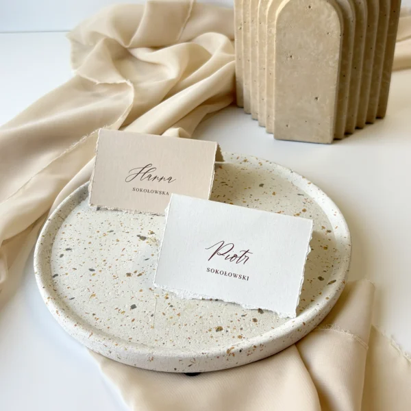 Dwie winietki w formie daszka – jedna w kolorze białym, druga beżowa – ustawione obok siebie na talerzu dekoracyjnym.