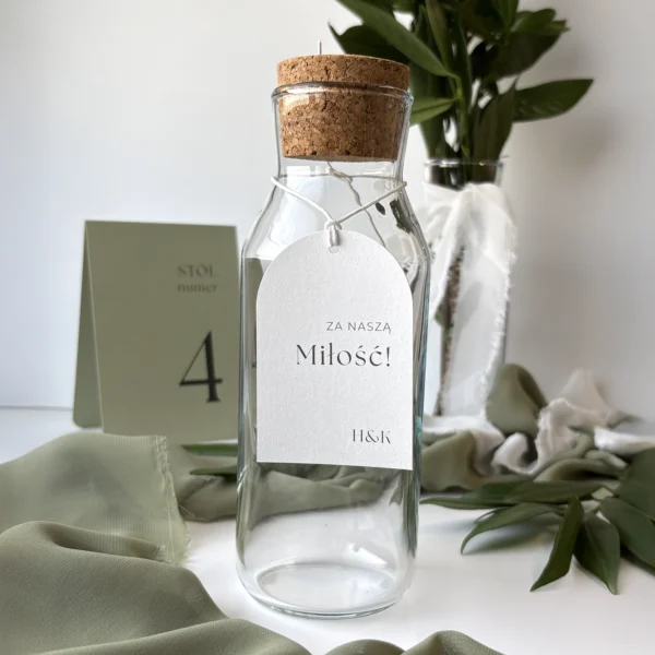 Zawieszka na alkohol Jungle w kolorze złamanej bieli z napisem „Za naszą miłość!”, zawieszona na sznureczku na szklanej butelce.