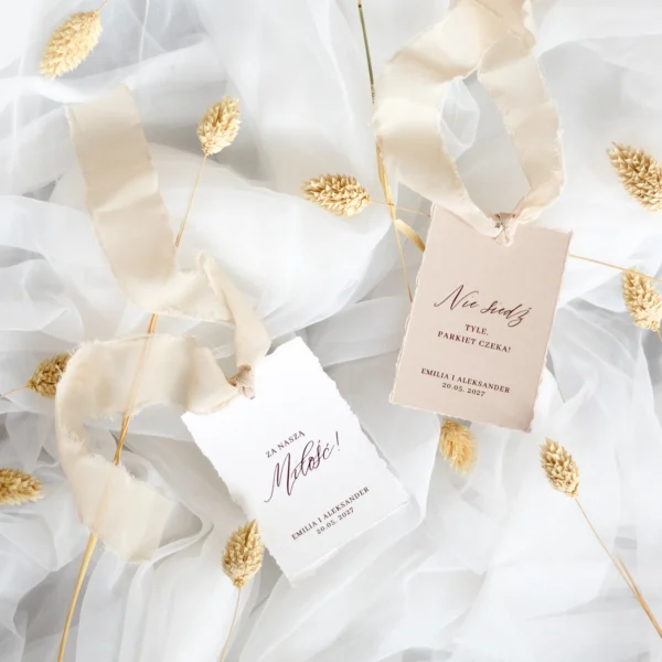 Zestaw zawieszek na alkohol z kolekcji Róża z beżową tasiemką, z napisami "Za naszą miłość!" oraz "Nie siedź, tyle parkiet czeka!", na tle jasnego tiulu i kłosów.