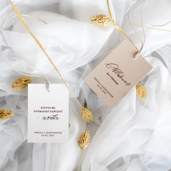 Dwie zawieszki na alkohol z kolekcji Róża – biała i beżowa – ze sznurkiem i zabawnymi tekstami, sfotografowane na tiulowym tle z dodatkiem kłosów.