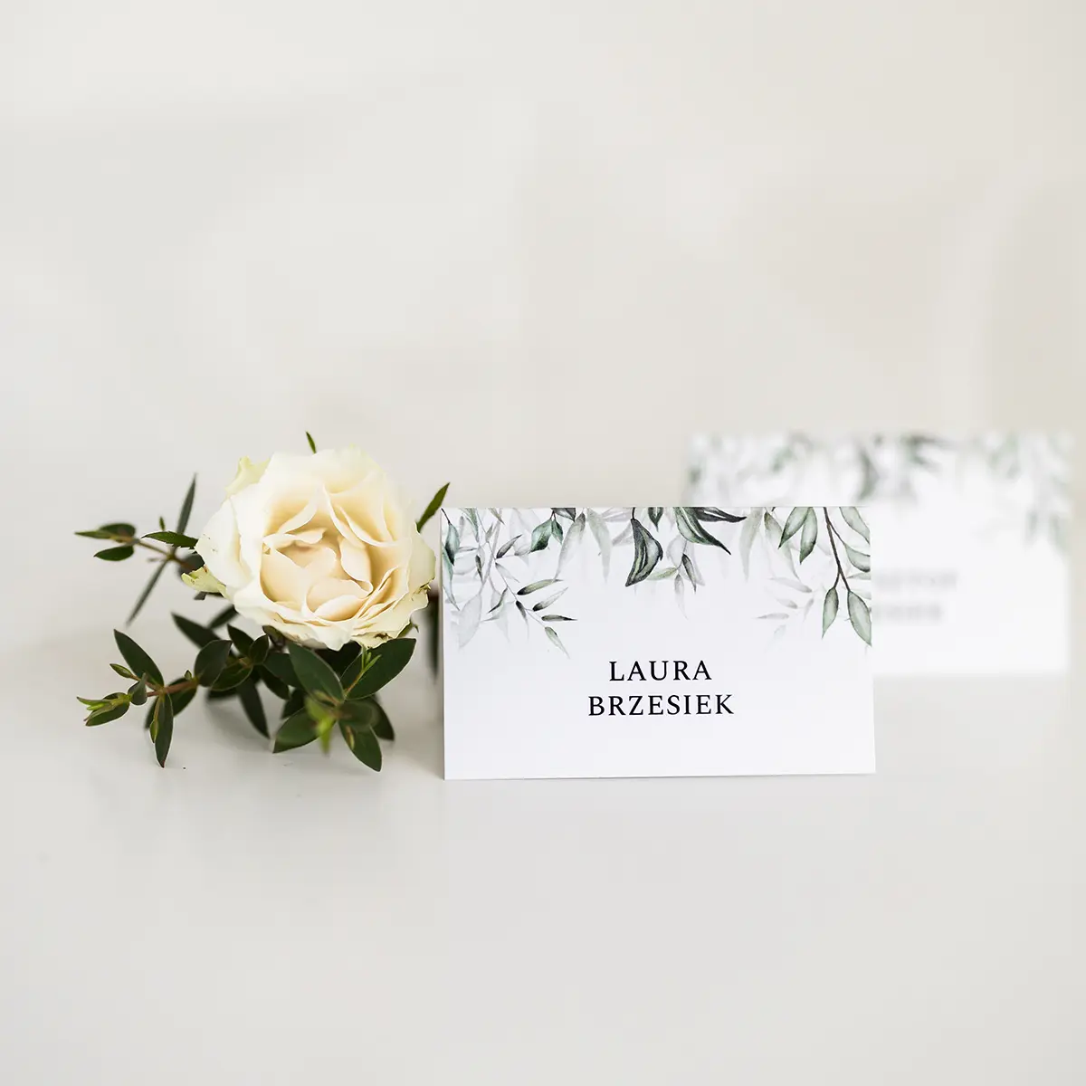 Pojedyncza winietka weselna z roślinnym wzorem w odcieniach zieleni, postawiona obok białej róży na neutralnym tle.