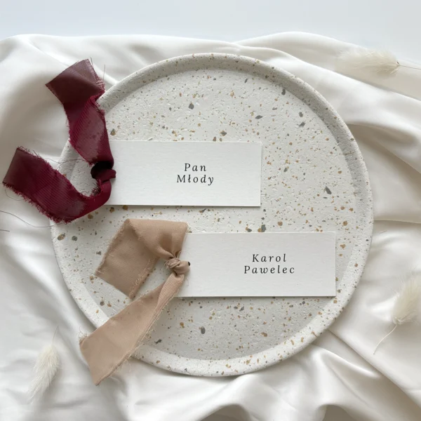 Winietki ślubne z bordową i beżową szarpaną wstążką, ułożone na talerzu z kamionki na tle satynowej tkaniny.