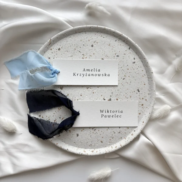 Winietki ślubne z granatową i błękitną szarpaną wstążką, prezentowane na jasnym tle z dekoracyjnym talerzem.