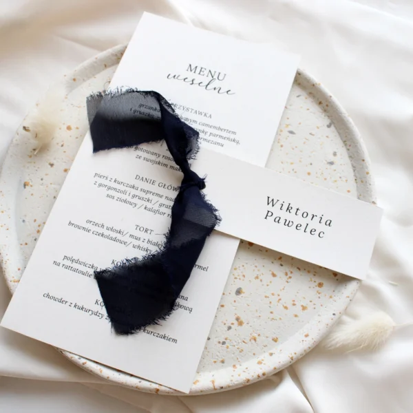 Stylowe menu weselne z granatową winietką przewiązaną szarpaną wstążką, ułożone na jasnym tle z subtelnymi akcentami.