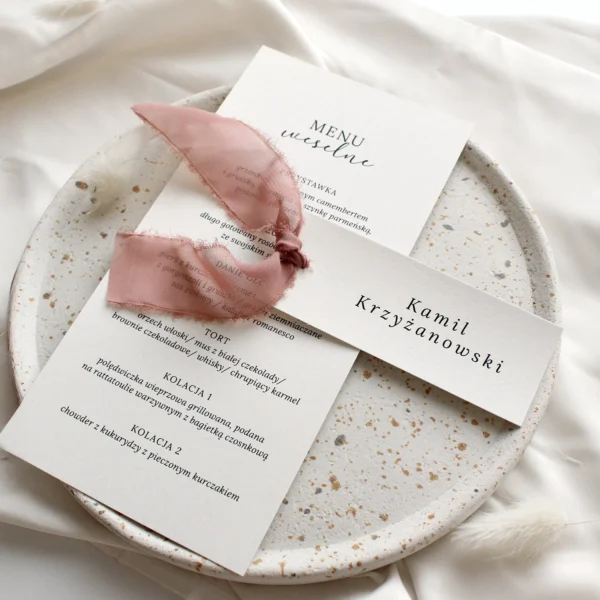 Menu weselne w eleganckim stylu z winietką ozdobioną różową, szarpaną wstążką, ułożone na talerzu z kamionki.