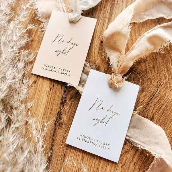 Dwie zawieszki „Na drugą nóżkę!” w kolorach białym i beżowym z szyfonowymi wstążkami na tle drewna.