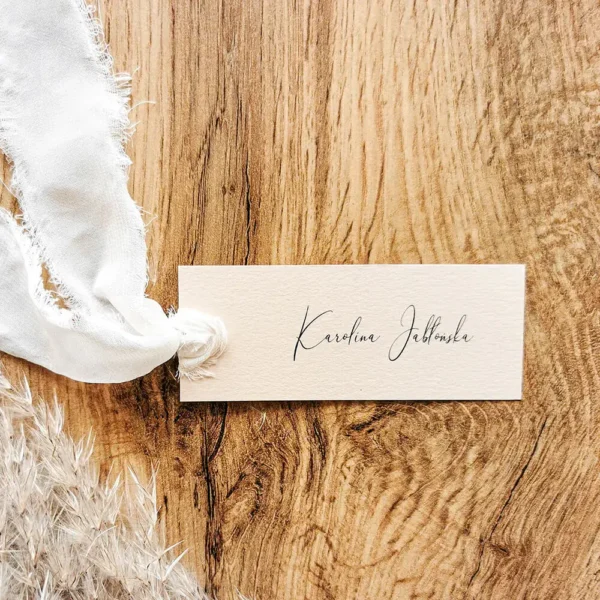 Beżowa winietka z imieniem gościa przewiązana białą szyfonową wstążką, ułożona na drewnianej powierzchni.