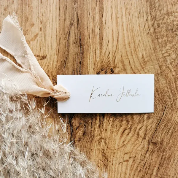 Biała winietka z eleganckim napisem, przewiązana beżową szyfonową wstążką na tle drewna i suszonych traw.
