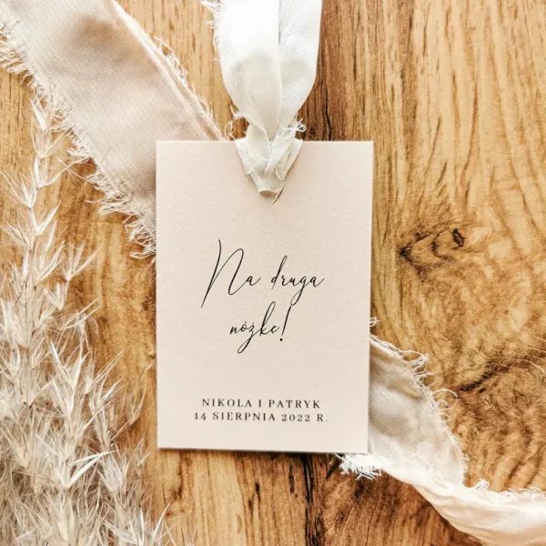 Beżowa zawieszka z napisem „Na drugą nóżkę!” przewiązana postrzępioną szyfonową wstążką, leżąca na drewnianym tle.