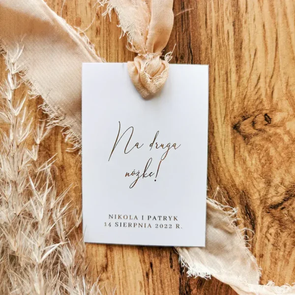 Biała zawieszka z eleganckim złotym napisem „Na drugą nóżkę!” i beżową szyfonową wstążką na drewnianym tle