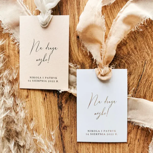 Komplet zawieszek „Na drugą nóżkę!” w wersji beżowej i białej z dekoracyjnymi postrzępionymi wstążkami, ułożone na drewnie.