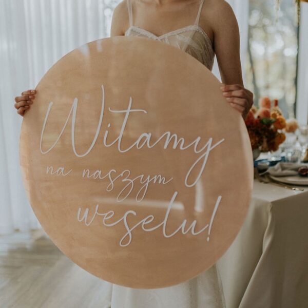 Okrągła tablica powitalna w kolorze beżowym trzymana prze pannę młodą.