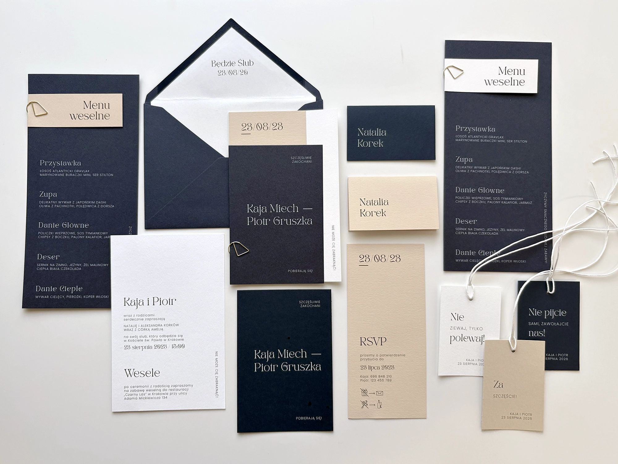 Zaproszenia ślubne z kolekcji Navy Blue w granatowo-beżowej kolorystyce z elegancką typografią i dodatkami, ułożone na jasnym tle.