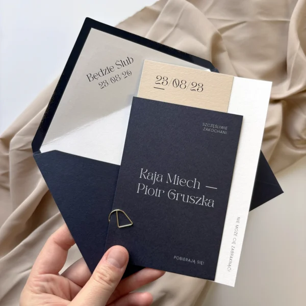 Granatowe zaproszenie ślubne z kolekcji Navy Blue z beżową i białą wkładką oraz ozdobną kopertą, trzymane w dłoni na tle beżowej tkaniny.