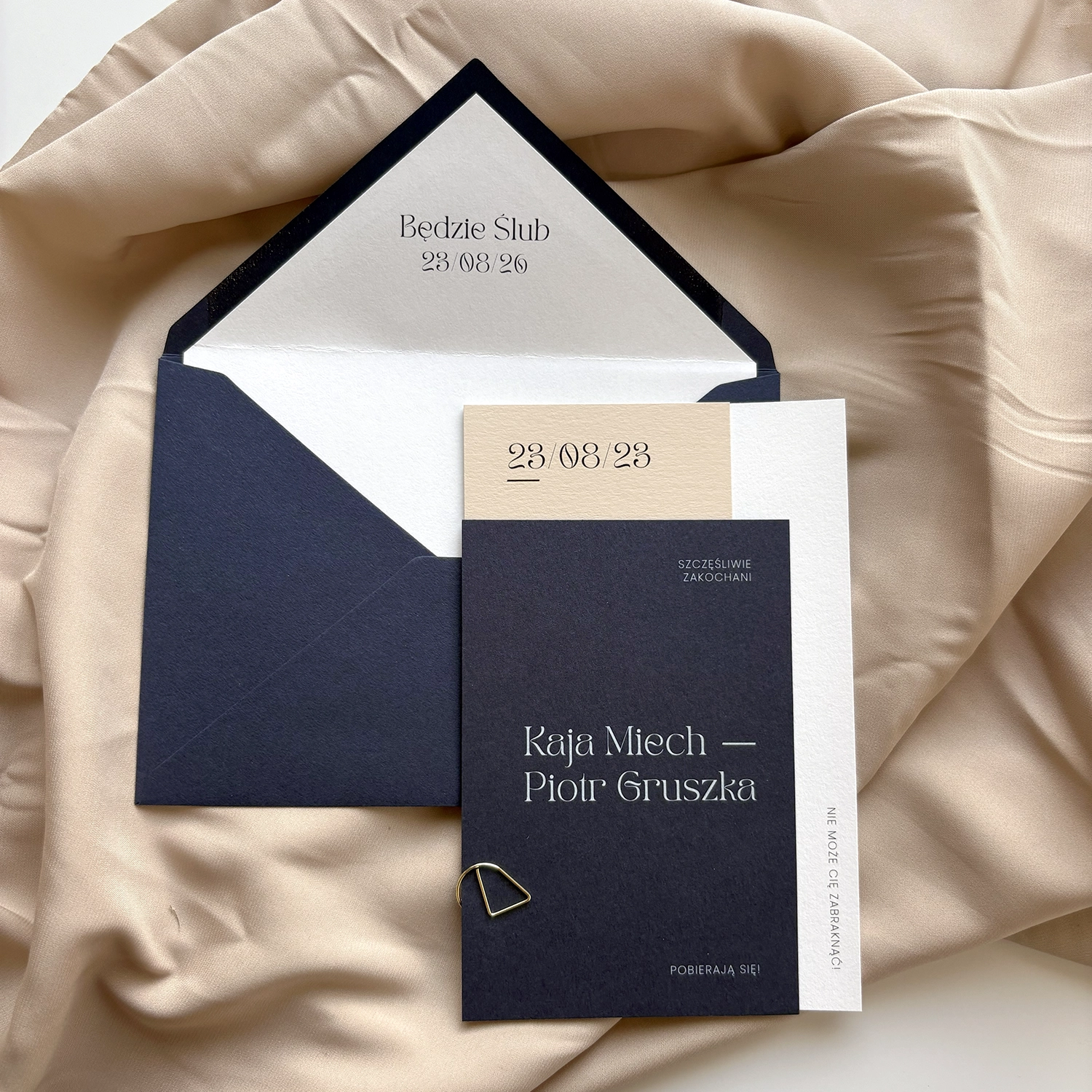 Eleganckie zaproszenia z kolekcji Navy Blue spięte złotym spinaczem, w tle tkanina w kolorze beżowym.