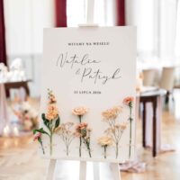 Kwiatowa tablica powitalna ustawiona na białej drewnianej sztaludze na tle sali weselnej