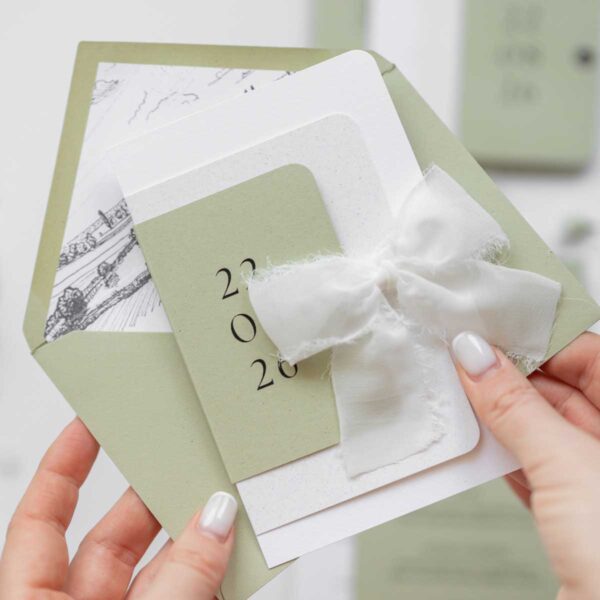 Zaproszenie weselne w kolorze oliwkowej zieleni i bieli z materiałową wstążką - Willa Słoneczna, trzymane w damskich dłoniach