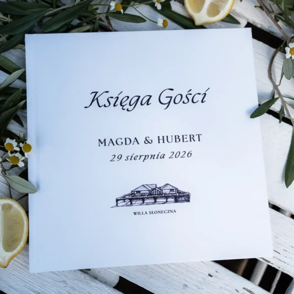 Księga gości z elegancką okładką, ozdobiona rysunkiem Willi Słoneczna oraz datą ślubu, położona na stole z dekoracjami.