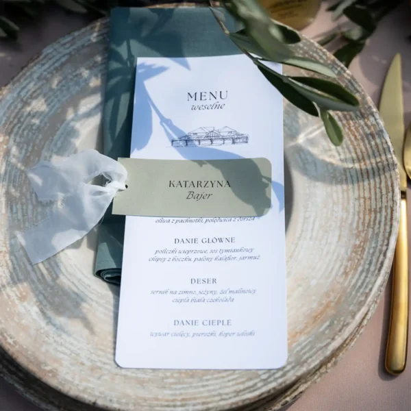 Stylowe menu weselne z eleganckim nadrukiem, leżące na talerzu w towarzystwie dekoracji oliwnych.