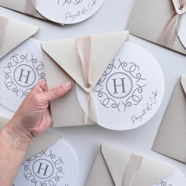 Elegancki, okrągły voucher podarunkowy z personalizowanym nadrukiem w delikatnej, beżowej kopercie z wstążką.