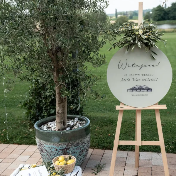 Tablica powitalna na wesele, ozdobiona kwiatami i liśćmi oliwnymi.
