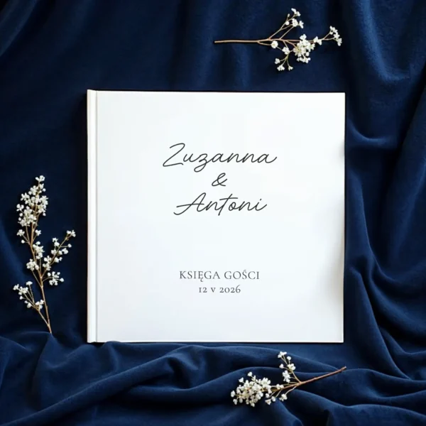 Biała księga gości z eleganckim napisem Zuzanna & Antoni, położona na granatowym materiale i ozdobiona gałązkami białych kwiatów.