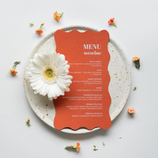 Leżące menu weselne z falowanymi krawędziami w kolorze pomarańczowym leżące obok kwiatka na talerzyku z wzorem lastryko
