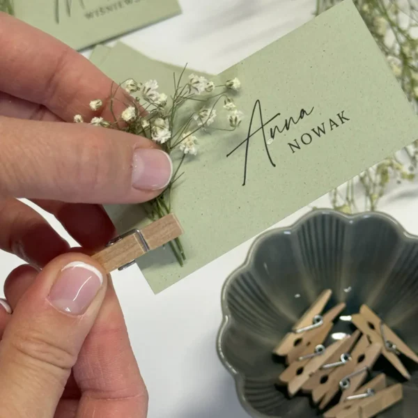 Osoba mocuje gałązkę suszonej gipsówki do winietki ślubnej w pastelowym odcieniu zieleni za pomocą małej drewnianej klamerki.