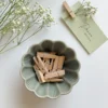 Mała miseczka ceramiczna z drewnianymi klamerkami, obok której leży winietka ślubna z przypiętą gipsówką, na jasnym tle.
