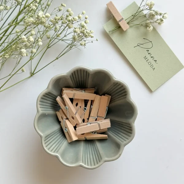 Mała miseczka ceramiczna z drewnianymi klamerkami, obok której leży winietka ślubna z przypiętą gipsówką, na jasnym tle.