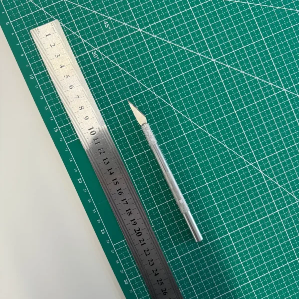 Zbliżenie na metalową linijkę i precyzyjny nożyk do cięcia leżące na macie do pracy ręcznej, używane do przygotowania winietek.