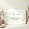 Elegancka tablica powitalna z delikatnym florystycznym motywem i kaligrafią, ustawiona w stylowej aranżacji.