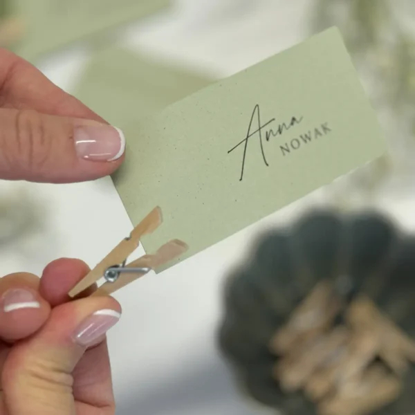 Ręka trzymająca winietkę ślubną w pastelowej zieleni, przypiętą drewnianą klamerką z dodatkiem suszonej gipsówki, na rozmytym tle dekoracji.