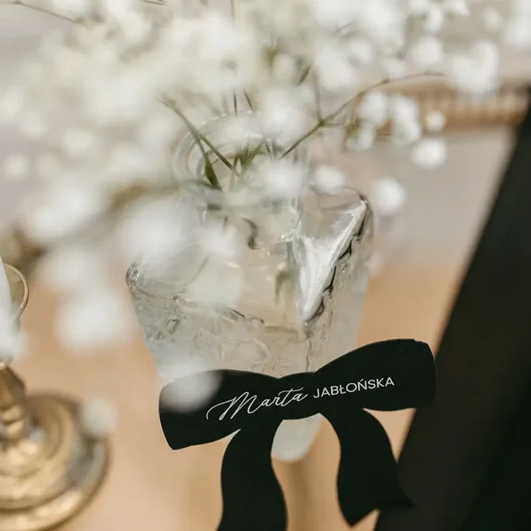 Winietka na stół w kształcie kokardki, pięknie komponująca się z delikatnymi dekoracjami kwiatowymi.