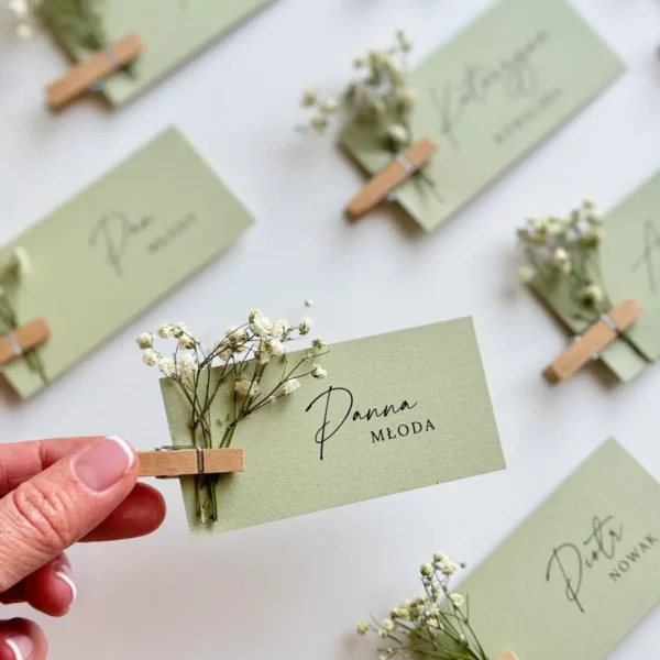 Delikatne zielone winietki ślubne DIY, każda przypięta drewnianą klamerką i ozdobiona suszoną gipsówką.