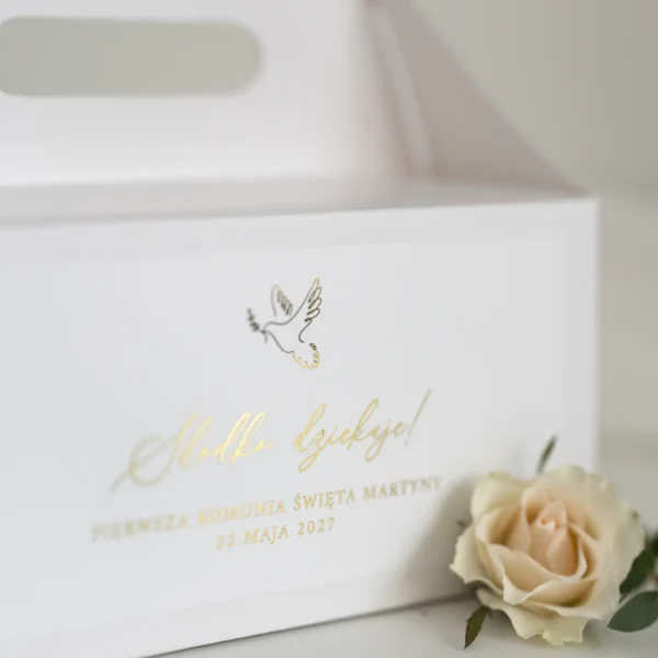Białe pudełko na ciasto z komunijną dedykacją w złotym kolorze oraz minimalistycznym wzorem gołębicy, obok kremowej róży na jasnym tle.