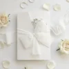 Eleganckie zaproszenie na komunię z gipsówką, przewiązane szyfonową wstążką, prezentowane w otoczeniu białych róż i płatków kwiatów.