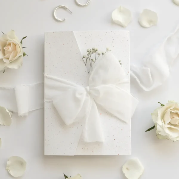 Eleganckie zaproszenie na komunię z gipsówką, przewiązane szyfonową wstążką, prezentowane w otoczeniu białych róż i płatków kwiatów.