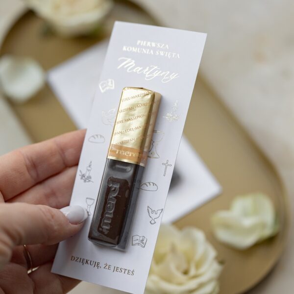 Osoba trzymająca w dłoni upominek z czekoladką Merci i kartą z napisem Pierwsza Komunia Święta Martyny oraz symbole religijne na tle złotej tacy.