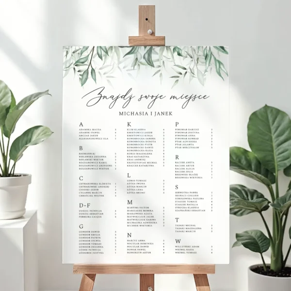 Alfabetyczny plan stołów na wesele z motywem zielonych liści, umieszczony na sztaludze, elegancki układ z czarną czcionką na białym tle.